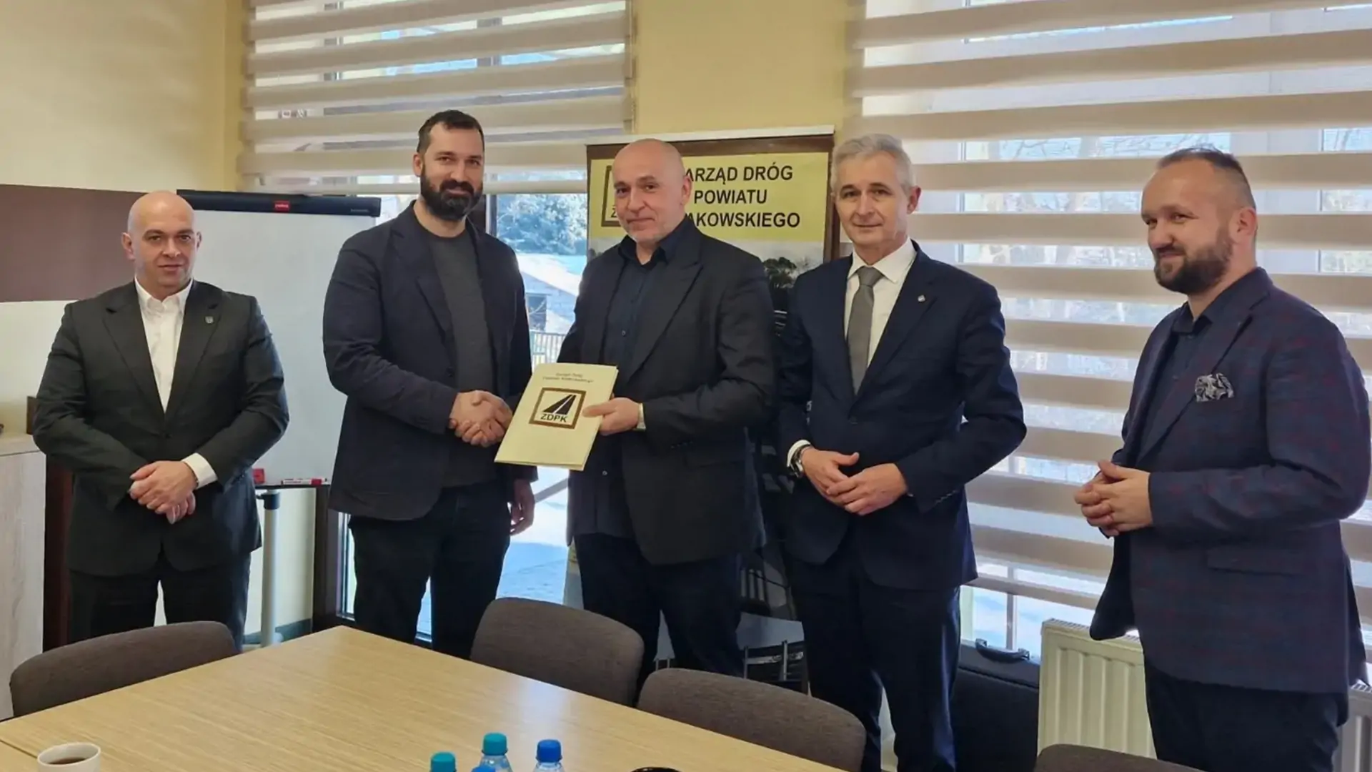 5 ubranych w garnitury mężczyzn pozuje do zdjęcia. Dwóch z nich trzyma umowę na przebudowę drogi. W tle roll-up Zarządu Dróg Powiatu Krakowskiego